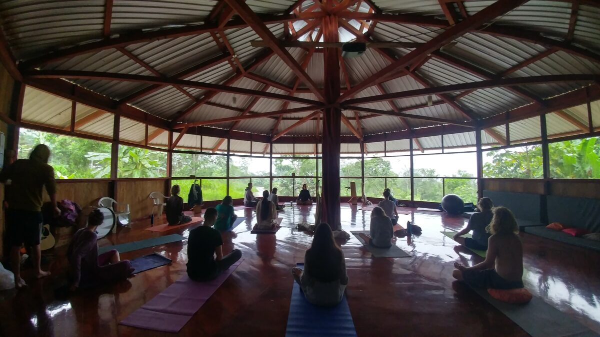 Photo of over a dozen people sitting cross-legged on yoga mats and pillows inside a wooden, octagonal structure with large screened-in windows looking-out at a lush green forest.
