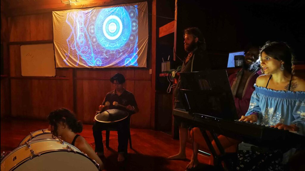 Photo of five people playing musi in a dark room. There is a projected image on the wall of some abstract visualizations.