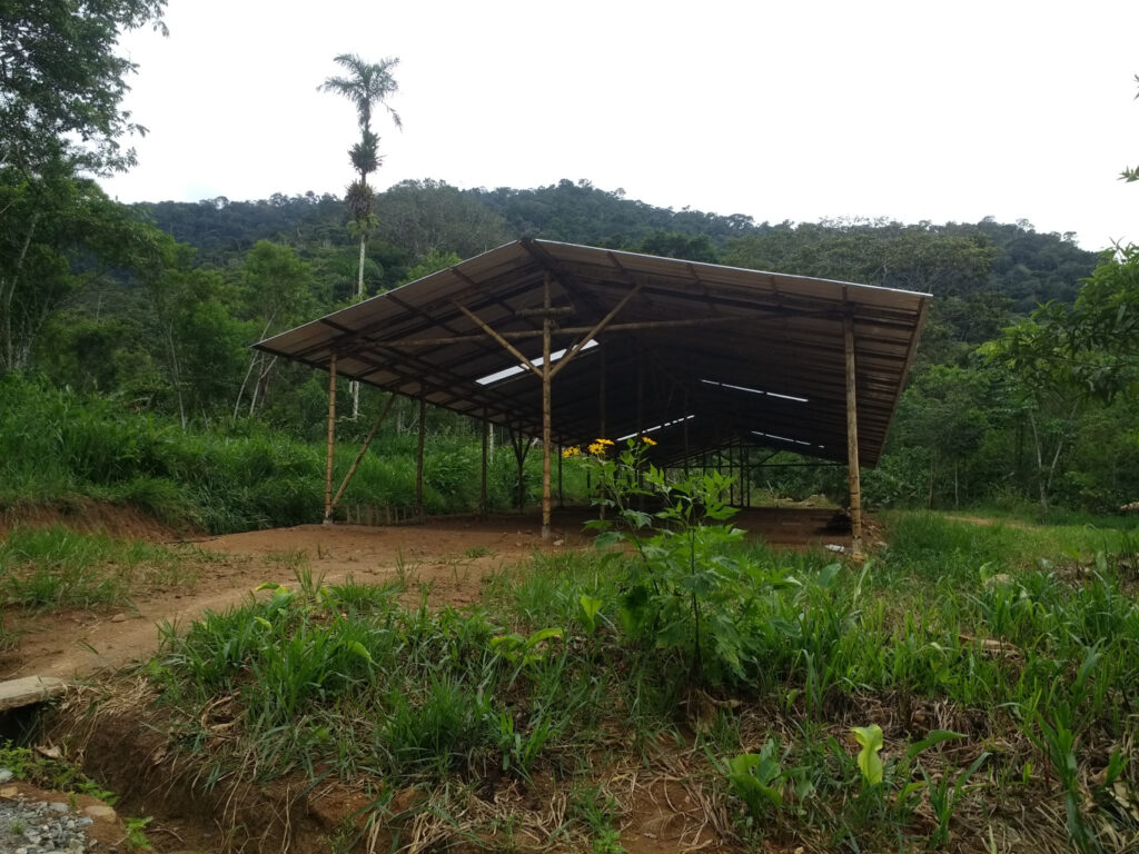 Photo of a large area with dirt floors covered by a simple roof structure. There is a small dirt path leading to the structure, and surrounding it is a lush green jungle.