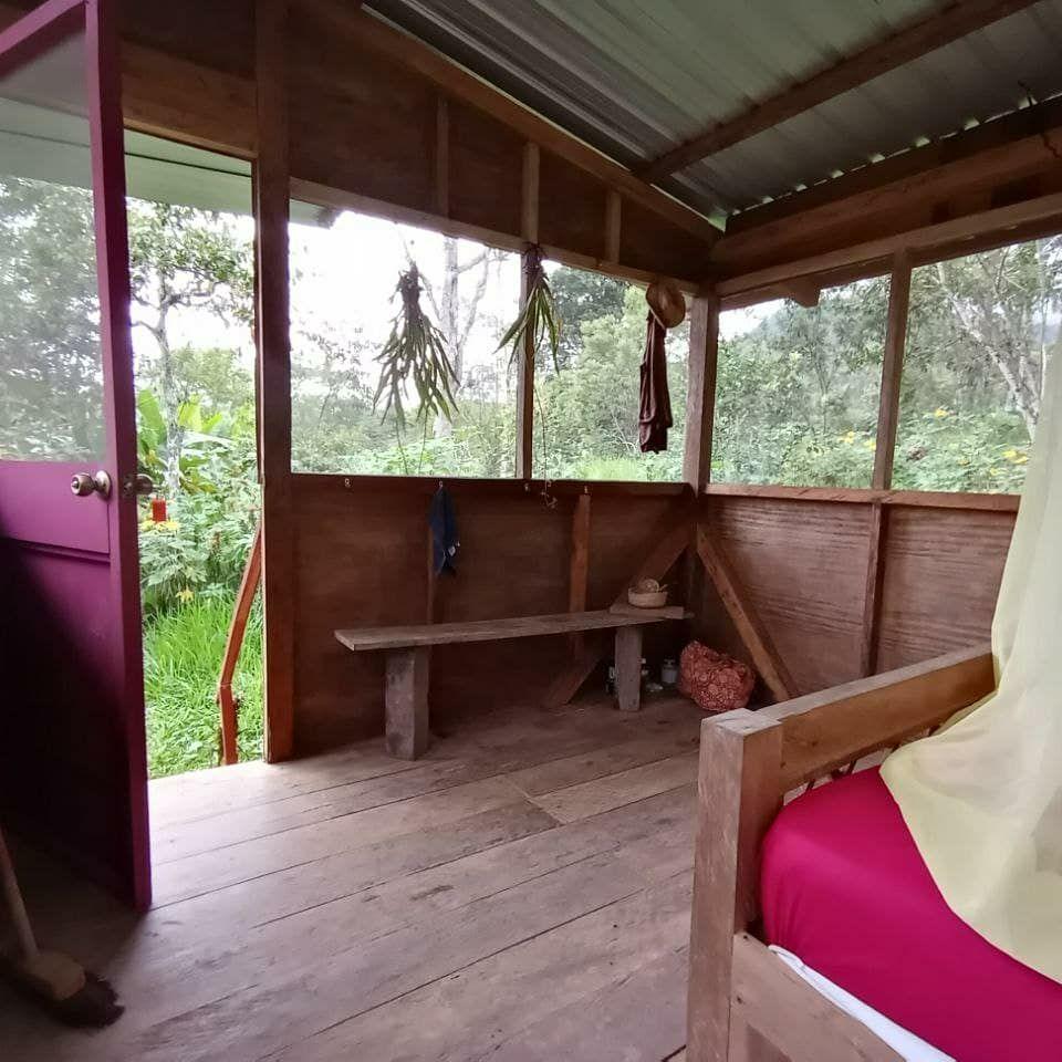 Photo inside a small, rustic bedroom with wooden floors and 360-degree screened-in windows. There is a small wooden bench and you can see the end of a wooden bed with a mattress and mosquito net. The door has a key in it, and there's a broom leaning against the wall.