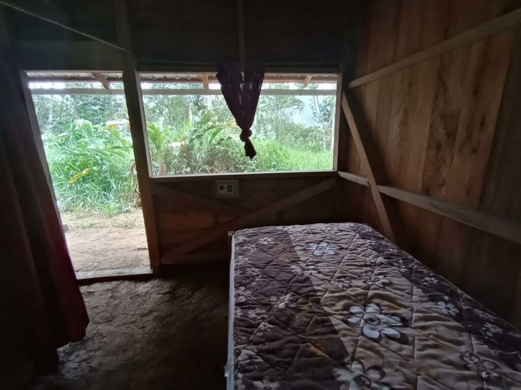 Photo of a small bedroom with wooden walls, a dirt floor, a bed with a mattress on it, and a small safe.