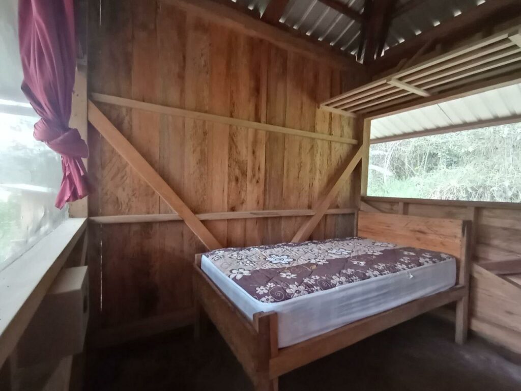 Photo of a small bedroom with wooden walls, a dirt floor, a bed with a mattress on it, and a small safe.