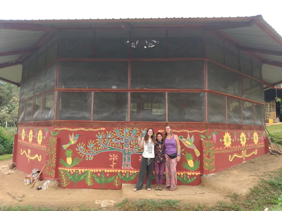 Photo of three people standing in-front of a structure artistically painted with a mural of trees, flowers, birds, pineapples, and other flora