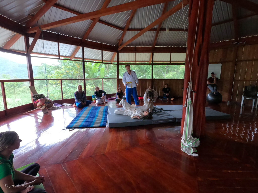 Two people practice jujitsu in the center of a circle of onlookers. One person is laying on a matt and another stands over them. An instructor stands next to them.