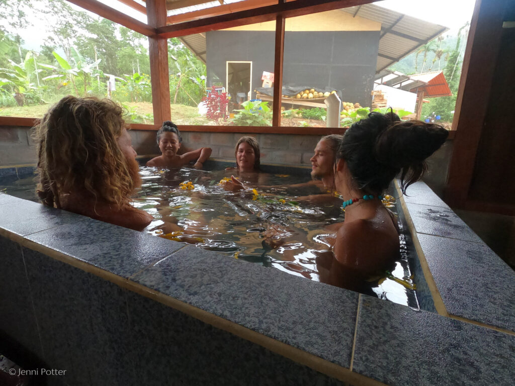 Photo of five people soaking in a large hot tub inside a screened-in structure. Through the large screened-in windows, we can see a structure, a large pile of coconuts, and lush jungle