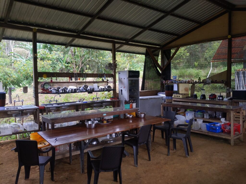 Photo of an outdoor kitchen with 3 sinks visible, a refrigerator, a chest freezer, a 4-burner gas stove, 3 blenders, 1 food processor, a lemon juicer, two large food prep areas, two water filters, compost buckets, seating for 5, and a wide variety of bowls and cutlery.