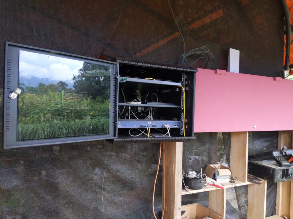 Photo shows an open rack with several pieces of server and networking equipment inside. A reflection of the glass cover of the rack shows the jungle with several trees, a row of lemongrass, yellow mexican sunflower, and bananas. An 18650 charger can be seen in the corner of the frame.