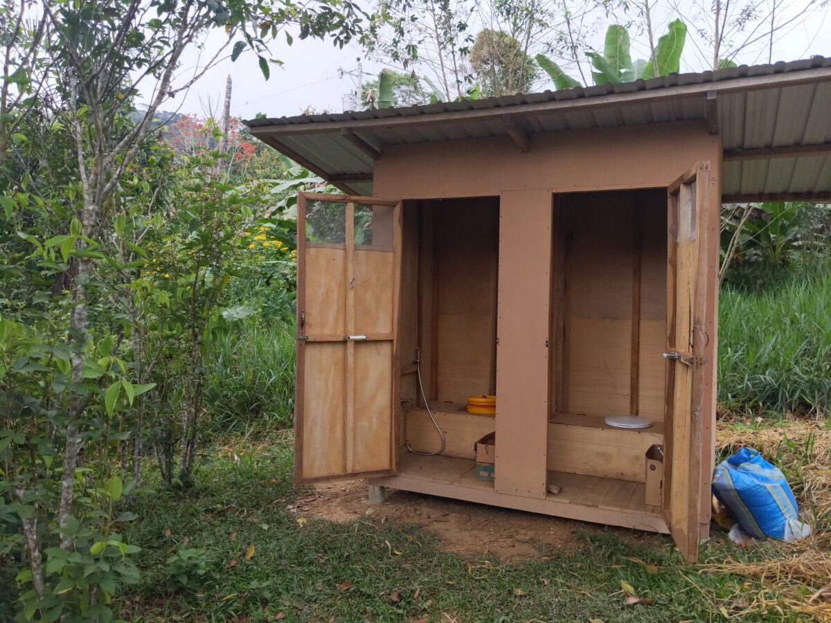 Photo of small structure with two doors open. Inside the right-door shows a toilet seat. Inside the left-door shows the top of a yellow bucket and a bidet hose mounted to the side of the wall. On the floor of each room is a large cardboard box.