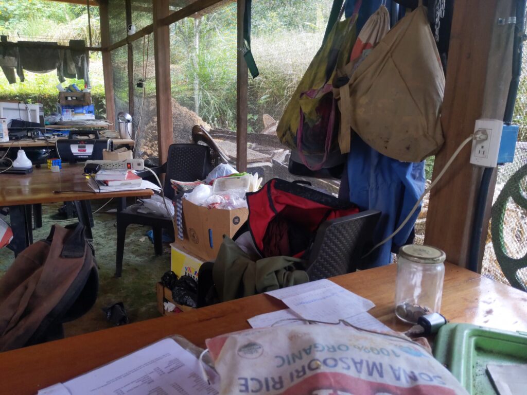 Photo of a table with a power outlet above it. The table behind has a power strip on it with a glowing red LED. The table behind that has several power cables going up to a power outlet that's dangling from above.