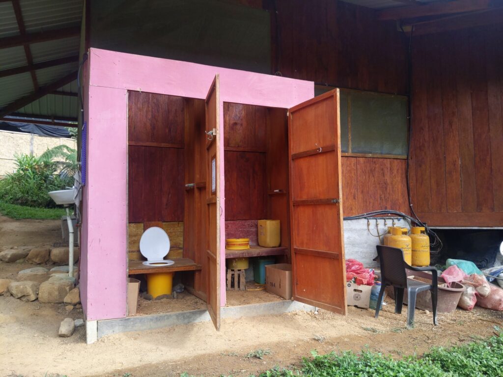 Photo of small structure with two doors open. Inside the right-door shows the top of a yellow bucket. Inside the left-door shows a toilet seat. On the floor of each room is a large cardboard box.