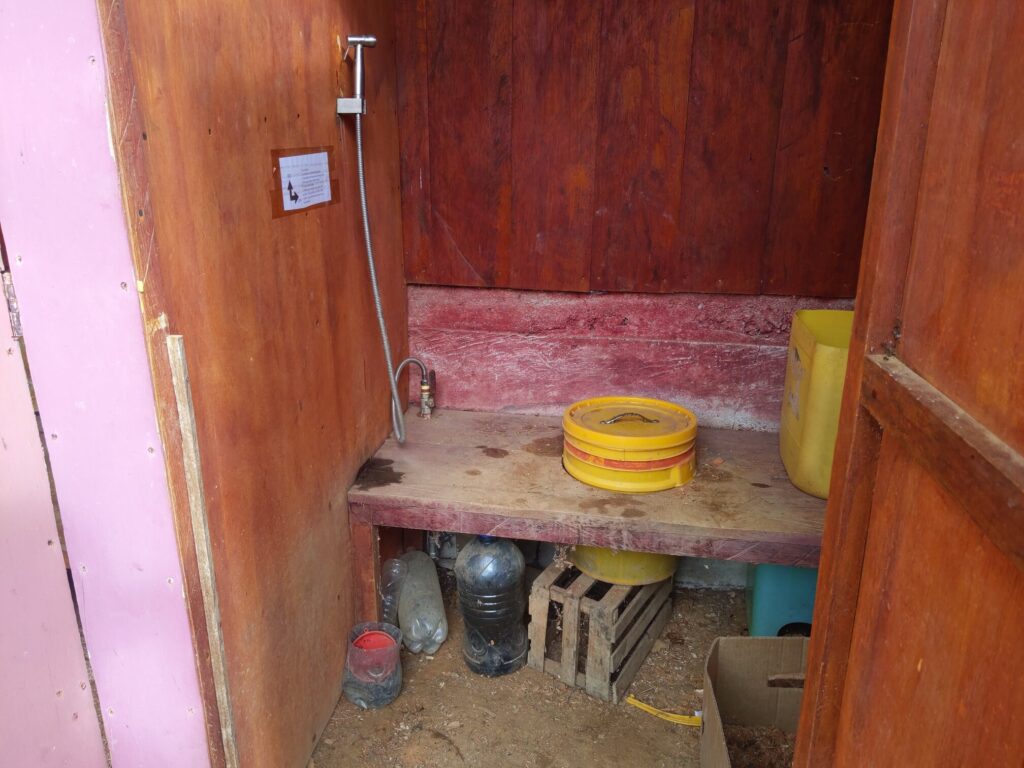 Photo of a small room with a yellow bucket mounted in a circular bench, supported from below by a wooden crate. To the left of the squat toilet is a bidet hose.
