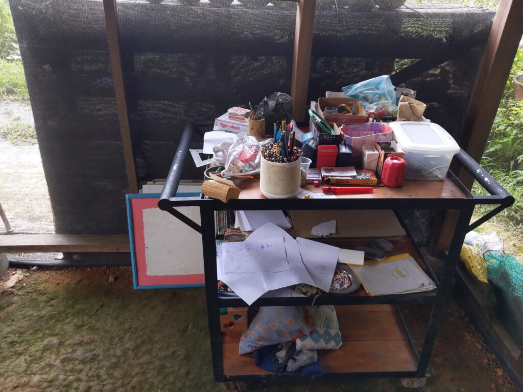 Photo shows a push cart with shelves cluttered with art supplies (colored pencils, pains, crayons, paper, chalk, sewing) and games (decks of cards and card games)